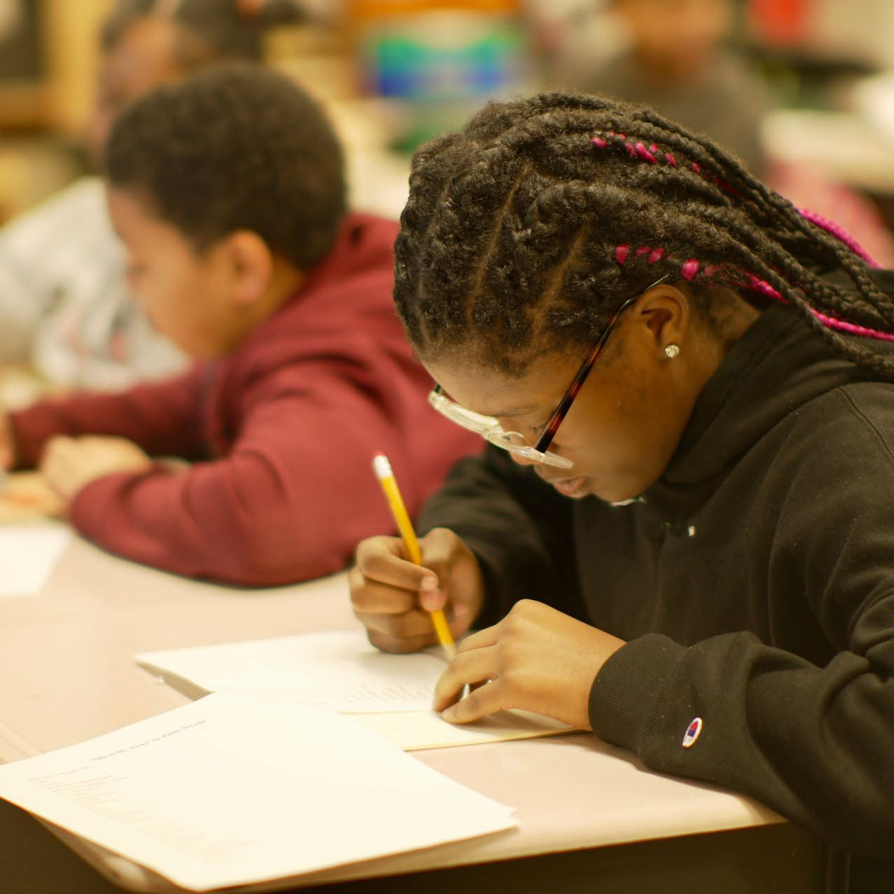 Academics Student wearing glasses writes on a classroom assignment while classmates work quietly in the background.